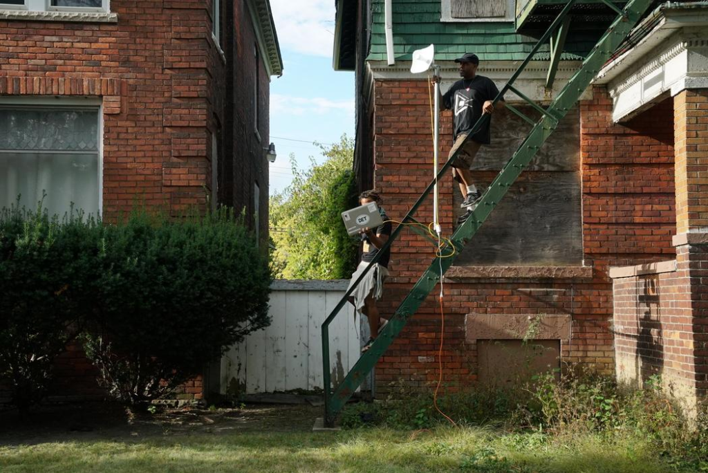 Two men walk down stairs connected to the exterior of a house. One holds a LAN network dish, the other holds a laptop. satellite dish from a house. 